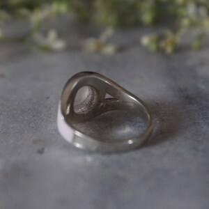 May include: A silver ring with a pink oval-shaped stone set in the center. The ring is on a gray surface.