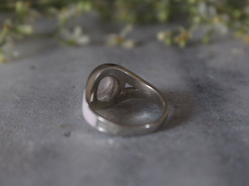 May include: A silver ring with a pink oval-shaped stone set in the center. The ring is on a gray surface.