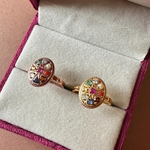 May include: Two gold rings with a circular arrangement of colourful gemstones, presented in a velvet jewellery box. The rings have a vintage aesthetic, featuring a central ruby and other gems.