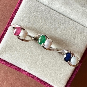 May include: Three silver rings, each with two oval gemstones. One ring features a ruby and a pearl, another has an emerald and a pearl, and the last one showcases a sapphire and a pearl. The rings are displayed in a pink velvet jewelry box.