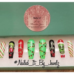 May include: A set of ten Christmas-themed press-on nails in a white box. The nails feature various designs, including candy canes, Christmas lights, and the Grinch. The box has a green lid with a sticker that says "Nailed It!" and "Thank You". The bottom of the box has the hashtag "#Nailed_It_By_Jewelz".