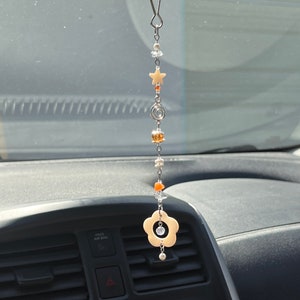 Peut inclure: Un charme en forme de fleur beige suspendu à une chaîne argentée avec un clip argenté. La chaîne est attachée au rétroviseur d'une voiture. Le charme a un cristal transparent au centre. La chaîne a également une étoile, une spirale et des perles orange et transparentes.