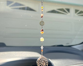 Puede incluir: Un adorno plateado para coche con un diseño de sol y luna. El adorno está ensartado con una cadena de cuentas naranjas y amarillas y un disco metálico decorativo. El adorno cuelga de un clip plateado.