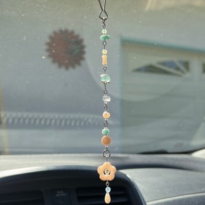 May include: A car charm with a flower-shaped pendant and a chain of beads in shades of green, beige, and white. The charm is hanging from the rearview mirror of a car.