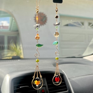 Puede incluir: Dos amuletos para coche colgados de un espejo retrovisor. Cada amuleto presenta una cadena dorada con cuentas, mariposas y una abeja o mariquita en un aro dorado. Los amuletos tienen detalles en amarillo, verde y rojo.