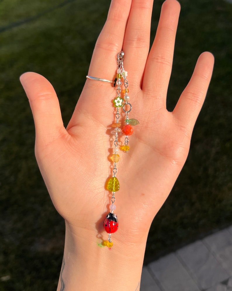 Pu&ograve; includere: Un braccialetto con ciondoli a catena d'argento con perline colorate, tra cui una coccinella, fiori e foglie.