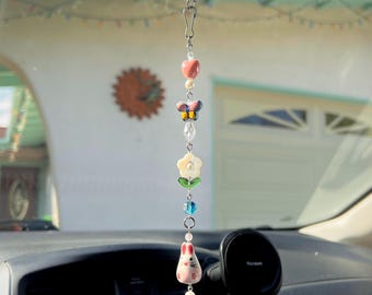 Pode incluir: Uma decoração de carro pendurada com um coração rosa, uma borboleta, uma flor e um amuleto de coelho com uma corrente prateada.