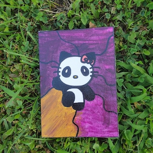 May include: A hand-painted canvas featuring a black and white Hello Kitty character with a pink bow on a purple and gold background.