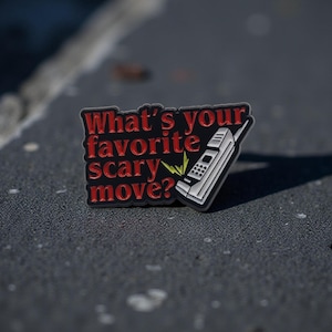Könnte beinhalten: Rechteckiger Anstecker mit dem Text "What's your favorite scary move?" in Rot und Weiß. Eine weiße und graue Retro-Handy-Grafik befindet sich auf der rechten Seite. Der Anstecker liegt auf einer grauen, strukturierten Oberfläche.