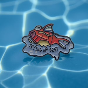 Peut inclure: Épingle en émail représentant un poisson de dessin animé rouge et jaune avec le texte "TRYING MY BEST" sur un fond bleu clair. L'épingle est posée sur un fond d'eau bleue avec des reflets de lumière.