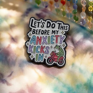 Könnte beinhalten: Ein schwarz-weißer Emaille-Pin mit dem Text "Let's Do This Before My Anxiety Kicks In." Der Pin zeigt einen grünen Frosch, der auf einem rosa Motorrad fährt. Der Pin ist vor einem Batik-Hintergrund platziert.