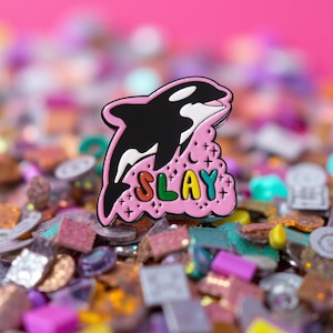 Könnte beinhalten: Ein Ansteck-Pin mit einem schwarz-weißen Orca, der über einer rosa Wolke springt, auf der das Wort "SLAY" in bunten Buchstaben steht. Der Pin befindet sich vor einem Hintergrund aus bunten Konfetti und einem hellrosa Hintergrund.