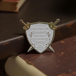 Könnte beinhalten: Weiß-goldener Emaille-Pin in Schildform mit zwei gekreuzten Schwertern. Der Pin zeigt den Text "DON'T TELL ME. WOMEN ARE NOT THE STUFF OF HEROES" umgeben von floralen Details.