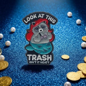 Könnte beinhalten: Emaille-Anstecknadel mit einer Waschbär-Meerjungfrau mit roten Haaren und einem türkisfarbenen Schwanz. Die Nadel trägt die Aufschrift "LOOK AT THIS" über dem Waschbären und "TRASH ISN'T IT NEAT?" darunter. Goldmünzen und Perlen auf blauem Hintergrund.