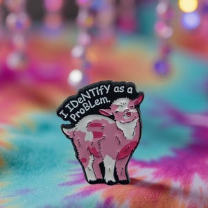 Könnte beinhalten: Ein rosa-weißes Ziegenabzeichen mit dem Text "I Identify as a Problem." Das Abzeichen hat einen schwarzen Rand und befindet sich auf einem bunten Batik-Hintergrund. Die Ziege lächelt.