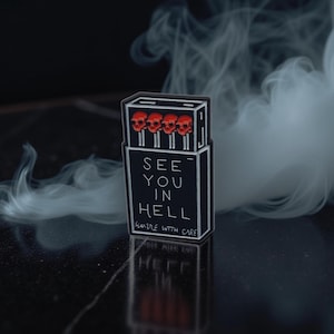 Può includere: Spilla nera a forma di scatola di fiammiferi con la scritta "SEE YOU IN HELL" e "HANDLE WITH CARE". I fiammiferi hanno teste di teschio rosse. La spilla è ambientata su uno sfondo scuro con volute di fumo bianco.