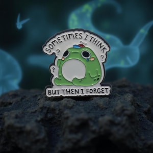 Könnte beinhalten: Ein grüner Frosch-Emaille-Pin mit dem Text "SOMETIMES I THINK BUT THEN I FORGET". Der Frosch hat einen traurigen Ausdruck mit Tränen und einem kleinen Hut. Der Pin ist weiß und schwarz mit dunklem Hintergrund.