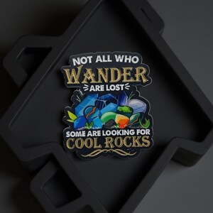 Könnte beinhalten: Ein Aufkleber mit dem Text "NOT ALL WHO WANDER ARE LOST" und "SOME ARE LOOKING FOR COOL ROCKS" über einer Grafik von bunten Steinen. Der Aufkleber befindet sich auf einem schwarzen, geometrischen Tablett.