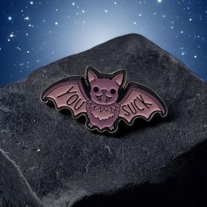 Könnte beinhalten: Ein lila fledermausförmiger Emaille-Pin mit den Worten "YOU SUCK" auf den Flügeln. Die Fledermaus hat ein lächelndes Gesicht. Der Pin befindet sich vor einem dunklen, strukturierten Hintergrund mit Sternenhimmel.