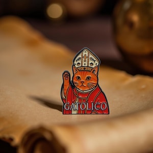 Könnte beinhalten: Ein farbenfroher Emaille-Pin mit einer orangefarbenen Katze, die als katholischer Papst gekleidet ist und ein rotes Gewand und eine Mitra trägt. Der Pin trägt das Wort "GATOLICO" in weißen Buchstaben. Der Pin ist vor einer Schriftrolle platziert.