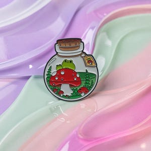 Könnte beinhalten: Emaille-Anstecknadel mit einem Frosch auf einem roten Pilz in einer Glasflasche. Die Flasche hat einen Korken und ein kleines Etikett. Der Hintergrund ist farbenfroh und gewirbelt.