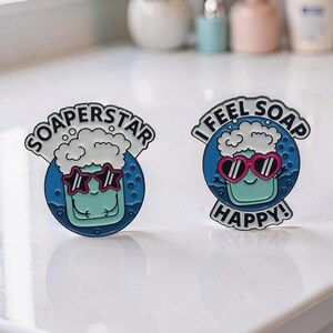 Könnte beinhalten: Zwei Emaille-Anstecknadeln mit Seifenthema. Eine Anstecknadel sagt "SOAPERSTAR" und die andere "I FEEL SOAP HAPPY!". Jede Anstecknadel zeigt einen lächelnden Seifencharakter mit Sonnenbrille, mit blauem Hintergrund und weißen Blasen.