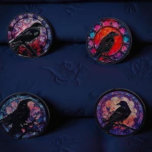 Może przedstawiać: Cztery okrągłe ozdobne przypinki z witrażowym wzorem przedstawiającym czarnego kruka siedzącego na gałęzi. Każda przypinka ma inną kolorystykę, w tym czerwoną, fioletową i pomarańczową, na ciemnoniebieskim tle.