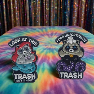 Könnte beinhalten: Zwei Emaille-Anstecknadeln mit Waschbär-Motiven. Eine zeigt einen Waschbären als Meerjungfrau mit dem Text "LOOK AT THIS" und "TRASH ISN'T IT NEAT?". Die andere zeigt einen Waschbären als Meerhexe mit "POOR UNFORTUNATE" und "TRASH".
