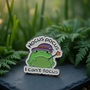 Puede incluir: Un pin de rana verde con un sombrero de bruja morado y una varita mágica. El pin tiene las palabras "Hocus Pocus" y "I can't focus" en texto blanco. Sobre una superficie gris oscura con plantas verdes de fondo.