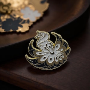 Könnte beinhalten: Ein goldfarbener Ansteckpin mit einem weißen Fuchsmotiv und einem floralen Schwanz. Der Pin hat schwarze und weiße Akzente und liegt auf einer dunklen Holzoberfläche. Der Pin ist ein dekoratives Accessoire.