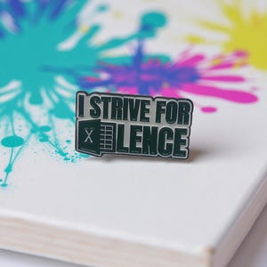 Puede incluir: Un pin rectangular de esmalte con el texto "I STRIVE FOR X LENCE" en letras blancas con un borde verde oscuro. El pin presenta un logotipo de Excel. El fondo es una superficie blanca con salpicaduras de pintura coloridas.