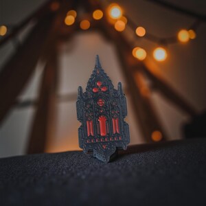 Könnte beinhalten: Ein schwarzer und roter Gothic-Pin in Form einer Kathedrale. Der Pin weist komplizierte Details auf, darunter gewölbte Fenster und dekorative Muster. Die roten Elemente heben sich vom schwarzen Hintergrund ab. Der Pin ist wahrscheinlich aus Metall oder einem ähnlichen Material.