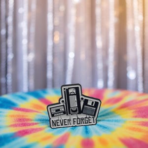 Puede incluir: Pin de esmalte en blanco y negro con un diseño de cinta de cassette, cinta de vídeo VHS y disquete. El pin tiene las palabras "NEVER FORGET" en fuente blanca. El pin está sobre un fondo tie-dye colorido.