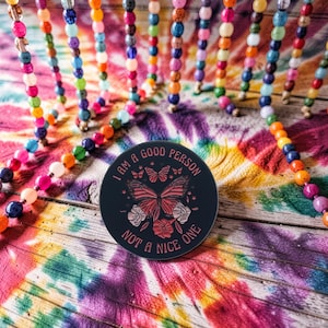 Könnte beinhalten: Ein runder, schwarzer Untersetzer mit dem Text "I AM A GOOD PERSON NOT A NICE ONE" und einem Schmetterlingsdesign. Der Untersetzer liegt auf einem Hintergrund mit Batikmuster und bunten Perlenschnüren.
