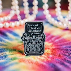 Könnte beinhalten: Ein schwarz-silberner Emaille-Pin in Form einer alten Schreibmaschine. Der Pin zeigt das Zitat "I am a writer. Therefore, I am not sane. -Edgar Allan Poe-" auf einem rechteckigen Feld. Der Hintergrund ist ein farbenfrohes Batikmuster.