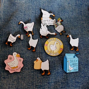 Peut inclure: Collection d'épingles en émail et d'une boîte de jus sur le thème de l'oie. Les épingles présentent des oies blanches avec des pieds et des becs orange dans diverses poses, certaines avec du texte comme "Silly Goose". La boîte de jus est bleue avec le texte "Silly Goose Juice 100%."