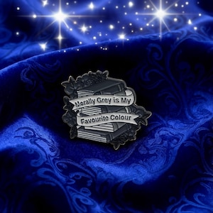 Könnte beinhalten: Ein Ansteck-Pin mit einem Stapel grauer und schwarzer Bücher und dem Text „Morally Grey is My Favourite Colour“ auf einem Banner. Der Pin ist mit floralen Elementen verziert, vor einem dunkelblauen, gemusterten Hintergrund.