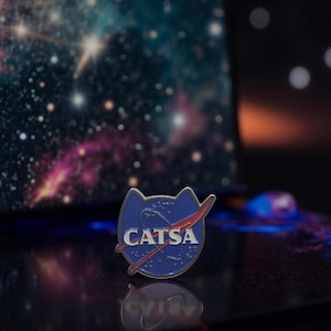 Könnte beinhalten: Blau-silberner Emaille-Anstecker in Form eines Katzenkopfes mit dem Wort "CATSA" in weißer Schrift. Der Anstecker zeigt ein rotes Swoosh-Design, das an das NASA-Logo erinnert, vor einem Weltraumhintergrund.