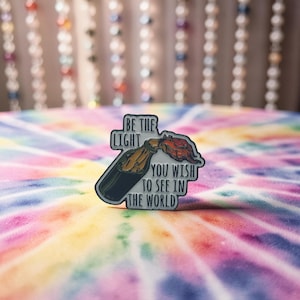Könnte beinhalten: Metall-Anstecknadel mit einem Flaschen- und Flammendesign und dem Text "BE THE LIGHT YOU WISH TO SEE IN THE WORLD". Die Nadel hat eine schwarze Umrandung, eine gold-braune Flasche und eine rote Flamme. Der Hintergrund ist ein Batikmuster.