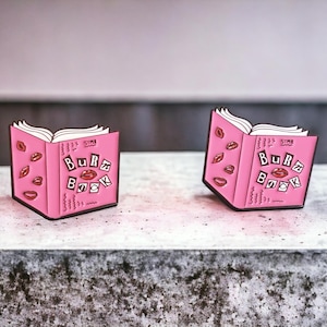 Könnte beinhalten: Zwei rosa, offen aussehende Buch-Anstecknadeln mit dem Text "Burn Book" und Lippenillustrationen. Die Anstecknadeln haben weiße Seiten und schwarze Umrisse. Eine Anstecknadel steht aufrecht, die andere ist leicht angewinkelt. Der Hintergrund ist ein neutrales Grau.