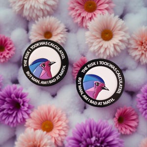 Könnte beinhalten: Zwei schwarze Emaille-Anstecknadeln mit einer Vogelillustration und dem Text "THE RISK I TOOK WAS CALCULATED, BUT MAN, AM I BAD AT MATH." Die Anstecknadeln sind von rosa, lila und pfirsichfarbenen Blumen umgeben.