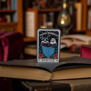 Könnte beinhalten: Ein rechteckiger Aufkleber mit dem Text "Trash Readers Book Club" zeigt einen Waschbären und einen Hamster, die in einem blauen Mülleimer lesen. Der Aufkleber hat einen schwarzen Rand mit floralen Akzenten und ist vor einem verschwommenen Hintergrund von Büchern platziert.
