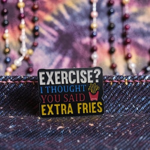 Könnte beinhalten: Schwarzer Emaille-Pin mit dem Text "EXERCISE? I THOUGHT YOU SAID EXTRA FRIES" in Weiß, Blau und Gelb. Der Pin zeigt eine rote Pommes-Grafik. Der Pin befindet sich auf einer Denim-Oberfläche.
