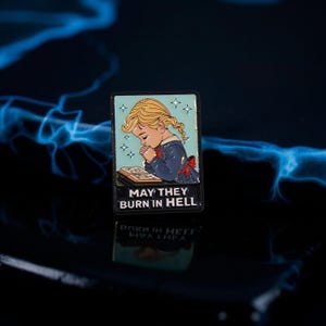 Puede incluir: Pin rectangular de esmalte con una ilustración retro de una niña rubia rezando sobre un libro. El pin tiene el texto "MAY THEY BURN IN HELL" en letras blancas sobre un fondo negro.