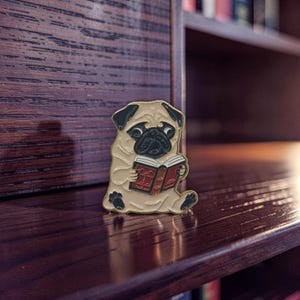 Può includere: Spilla smaltata raffigurante un carlino beige che legge un libro rosso. Il carlino ha dettagli neri sul viso e sulle orecchie. La spilla è ambientata su uno sfondo di legno scuro, che evoca un'atmosfera da biblioteca o studio.