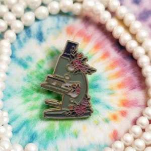 Könnte beinhalten: Dekorative Emaille-Anstecknadel in Form eines hellgrünen Mikroskops, verziert mit rosa und roten Blumen. Die Nadel befindet sich vor einem farbenfrohen Batik-Hintergrund und wird von einer Perlenkette umrahmt. Detailliertes Mikroskop-Design.