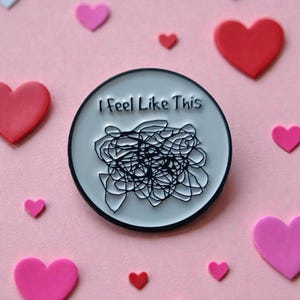 Könnte beinhalten: Runder Emaille-Anstecker mit dem Text "I Feel Like This" über einem schwarzen Kritzel-Design. Der Anstecker ist von rosa und roten herzförmigen Dekorationen auf rosa Hintergrund umgeben.