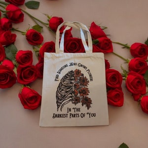 Könnte beinhalten: Weiße Einkaufstasche mit einer Schwarzweißillustration eines Brustkorbs, aus dem Blumen wachsen. Der Text auf der Tasche lautet "Finde jemanden, der Blumen in den dunkelsten Teilen von dir wachsen lässt."