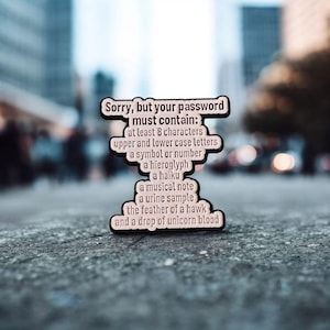 Pode incluir: Um alfinete de esmalte preto e branco com o texto "Sorry, but your password must contain:" seguido de uma lista de requisitos. O alfinete tem a forma de uma nuvem e está sobre um fundo urbano desfocado.