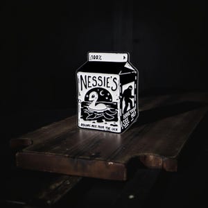 Pode incluir: Caixa de leite a preto e branco com o texto "Nessie's" e uma ilustração do Monstro do Lago Ness. A caixa também apresenta o texto "Organic Milk From The Loch" e "Have You Seen Me?"